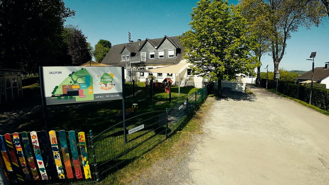 Blick vom Zufahrtsweg auf die St. Elisabeth KiTa Schönau, links der bunt bemalte Zaun und die Lageplan-Tafel